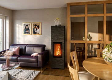 Frontalansicht eines schlanken, dunklen Naturstein-Kaminofens vor einer hellen Wand neben einem violetten Sofa.