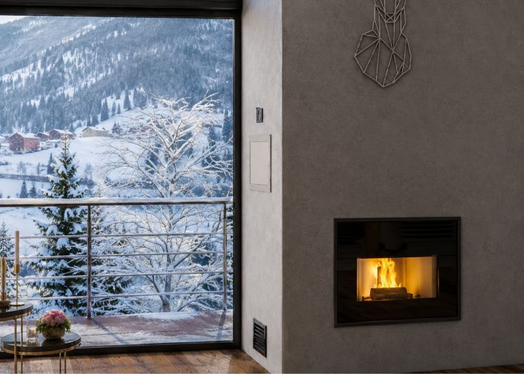 Nahaufnahme der JØTUL PF 901 Pelletkassette, die bündig in die graue Wandstruktur integriert ist, neben einer Balkontür mit Winterlandschaft.