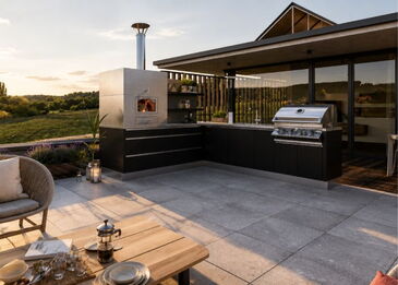Gesamtansicht der L-förmigen Edelstahl-Außenküche in Schwarz mit integriertem gemauerten Pizzaofen und Gasgrill bei Sonnenuntergang.