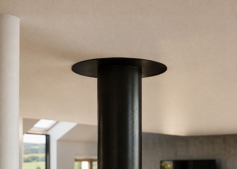 Detaillierte Ansicht der minimalistischen, runden Deckenplatte und des schwarzen Abgasrohrs, an dem der Kamin frei im Raum schwebt.