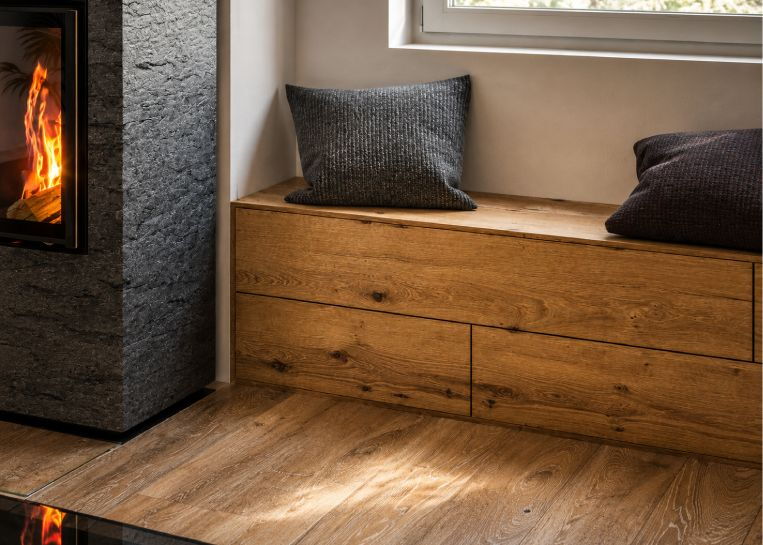 Detaillierte Ansicht der ergonomischen Holz-Sitzbank mit integrierten Schubladen, die direkt an den Kaminofen anschließt.