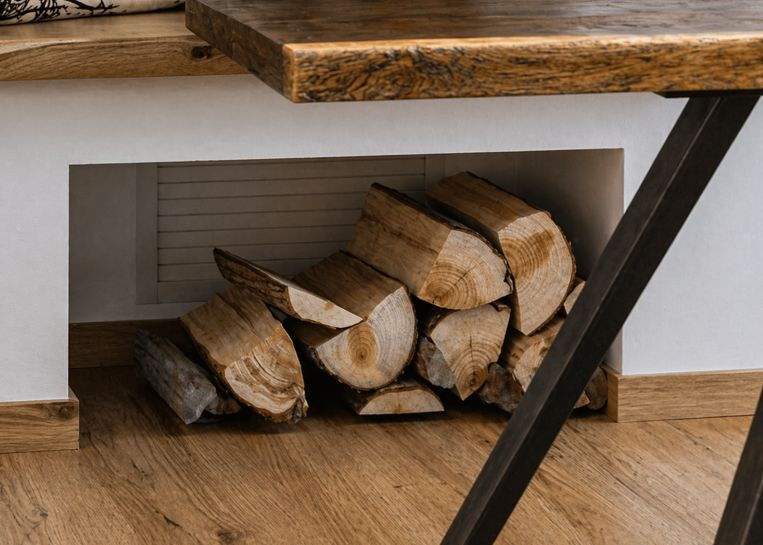 Nahaufnahme eines unter der Kaminsitzbank integrierten Holzfaches, in dem gespaltene Holzscheite griffbereit und dekorativ lagern.