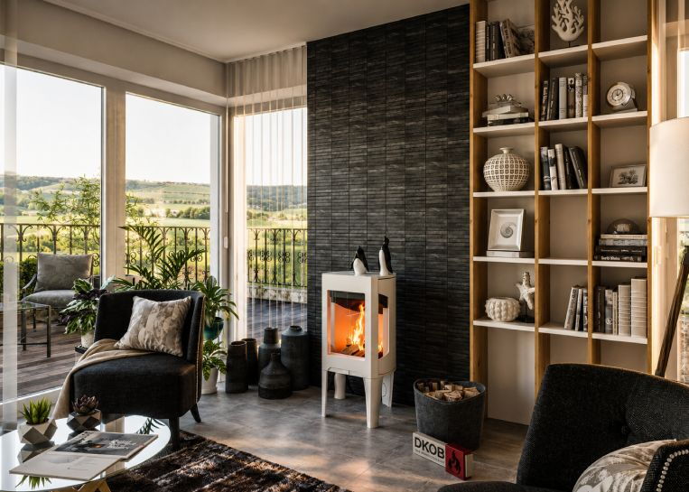Gesamtansicht des weißen Jotul F163 Kaminofens vor einer dunklen Natursteinwand in einem hellen Wohnzimmer mit Balkonausblick.