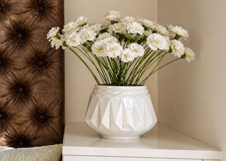 Eine geometrisch geformte, weiße Blumenvase mit frischen Nelken auf einem Sideboard neben der gemütlichen Kamin-Sitzbank.