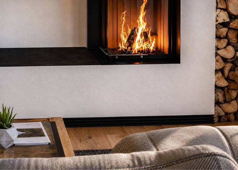 Nahaufnahme des lodernden Feuers im Brunner Eckkamin mit Fokus auf die moderne, schwarz gestufte Sockelleiste am Boden.