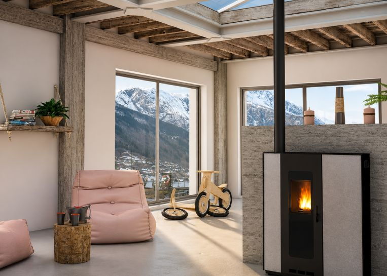 Ein flacher JØTUL PF 710 Pelletofen mit grauer Keramikverkleidung in einem hellen Zimmer mit Bergpanorama und Holzbalkendecke.