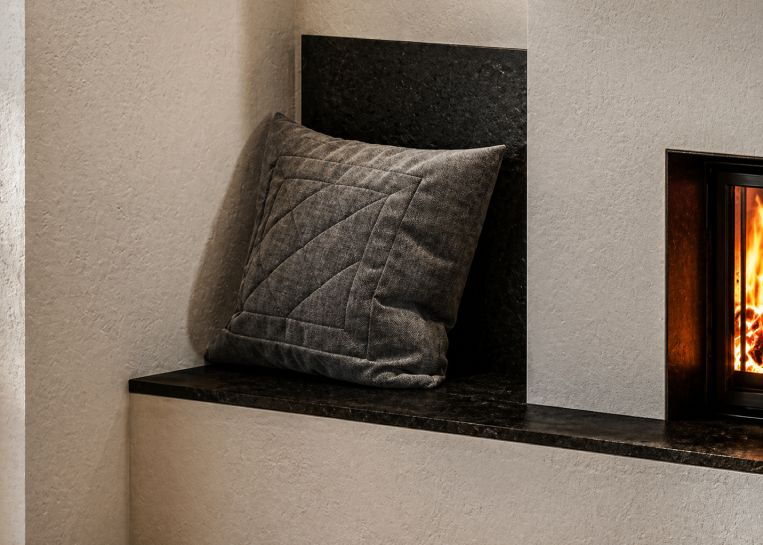 Nahaufnahme der gemütlichen Sitzbank am Kamin mit einer Rückenlehne aus dunklem Nero Assoluto Naturstein und einem dekorativen Kissen.