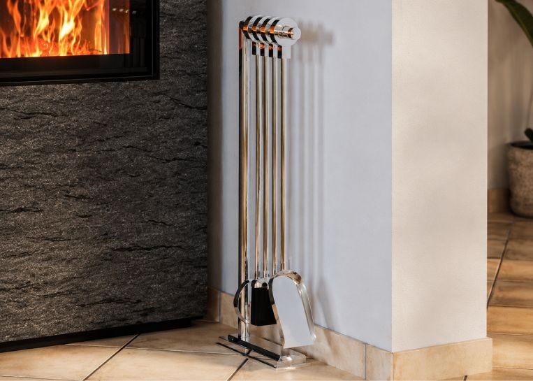 Nahaufnahme eines vierteiligen, hochglänzenden Kaminbestecks neben der strukturierten Natursteinwand des Ofens.