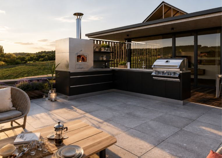 Gesamtansicht der L-förmigen Edelstahl-Außenküche in Schwarz mit integriertem gemauerten Pizzaofen und Gasgrill bei Sonnenuntergang.