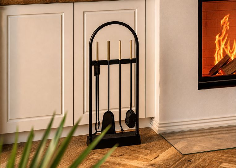 Ein elegantes Kaminbesteck in Schwarz-Gold steht neben der Kaminanlage auf dem Parkettboden mit transparenter Vorlegeglasplatte.