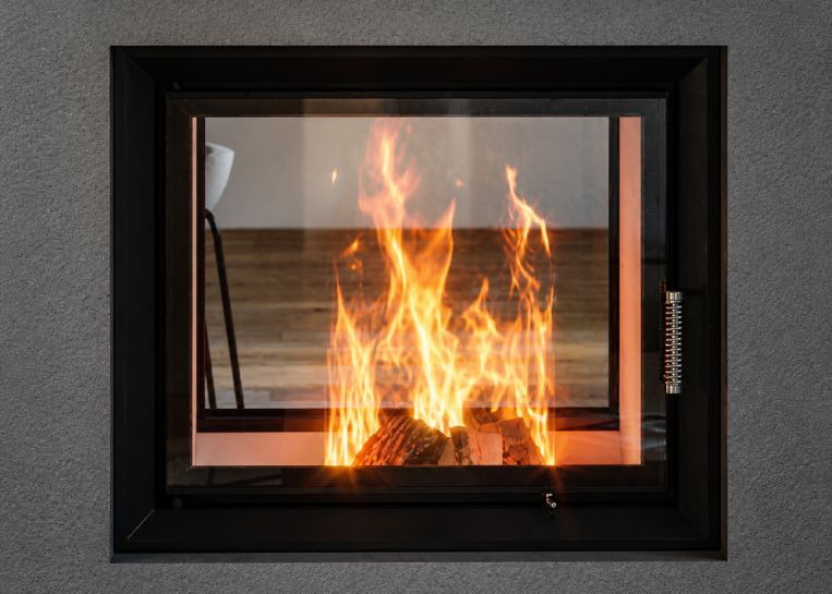 Nahaufnahme des lodernden Holzfeuers hinter der Sichtscheibe des Tunnelkamins, eingerahmt von der groben Struktur der Fresco-Spachtelmasse.