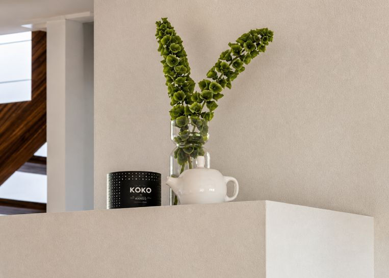 Detailansicht der glatt verputzten Kaminhülle mit einer eleganten weißen Vase, grünen Blumen und einer dekorativen Duftkerze.