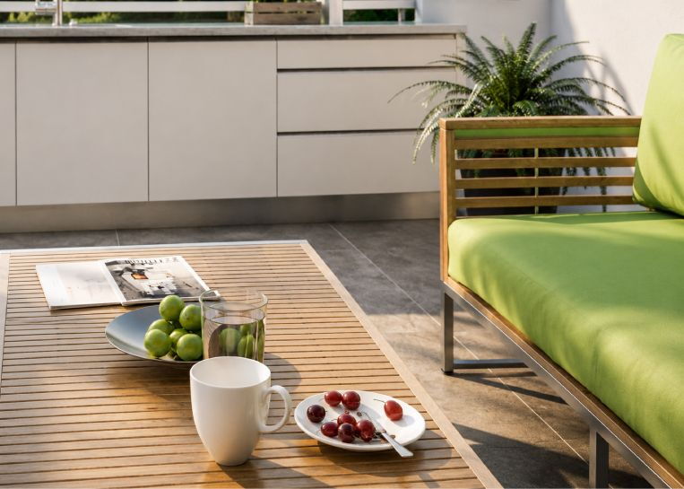 Nahaufnahme eines Holz-Loungetisches mit Obst und Getränken, im Hintergrund die weiße Edelstahl-Außenküche.