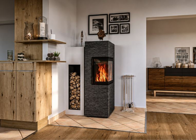 Frontale Ansicht des Kaminofens mit dunklem Naturstein und danebenliegendem gemauerten Körper inklusive Metallbox für Kaminholz.
