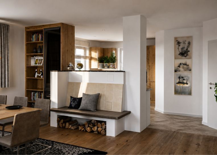 Ansicht der Rückseite der Herdanlage im Wohnbereich mit einer gemütlichen Sitzbank, wärmespeichernder Feinkeramik und integriertem Holzlager.