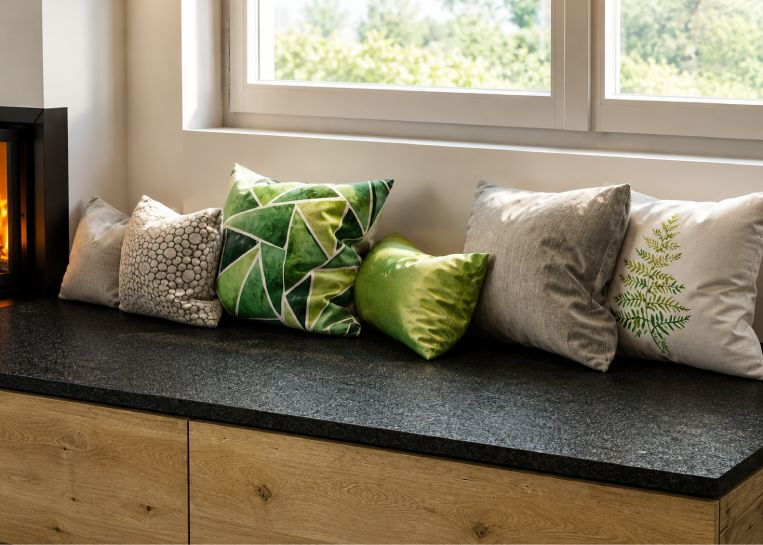 Detailansicht der speicherfähigen Sitzbank mit dunkler Natursteinplatte und dekorativen Kissen vor einem Fenster.